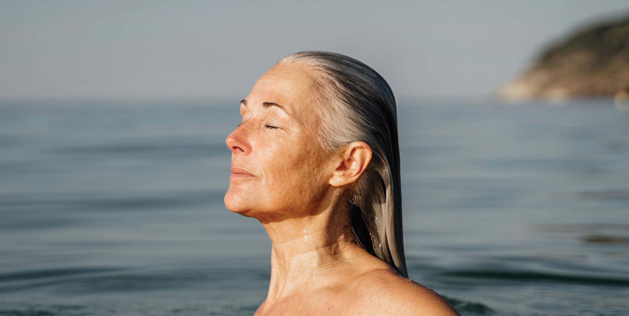 Elderly woman standing in the clean sea water with serene expression and eyes closed, enjoying the sunlight on her face.