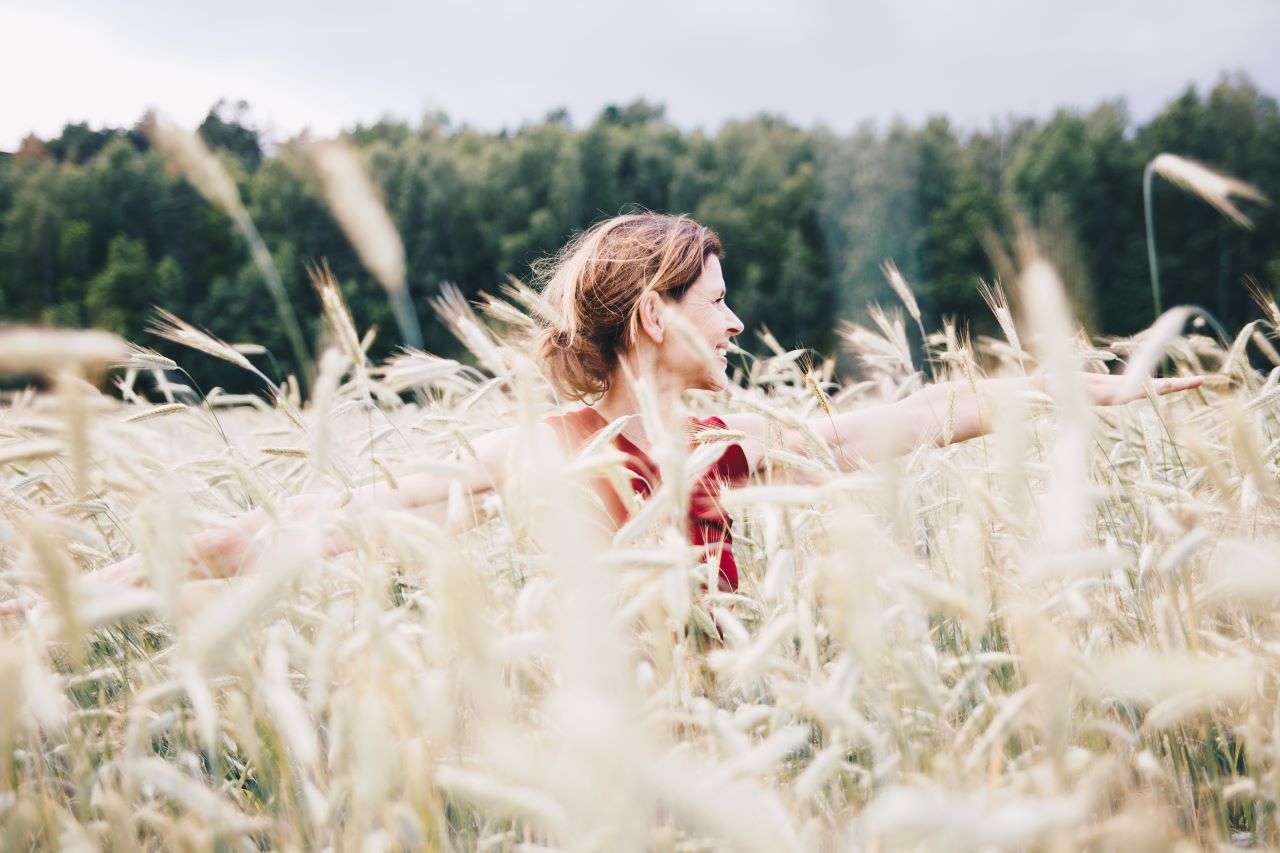 A smiling woman with arms outstretched in a wheat field with a forest in the background